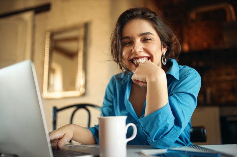 cheerful-attractive-young-woman-enjoying-distant-work-sitting-desk-using-portable-computer-drinking-coffee-pretty-female-blogger-working-from-home-uploading-video-her-channel-smiling (1)